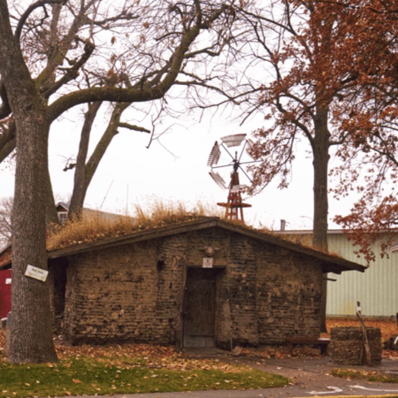 A small, stone building with a grass roof sits in the foreground, while a windmill and bare trees are visible in the background.