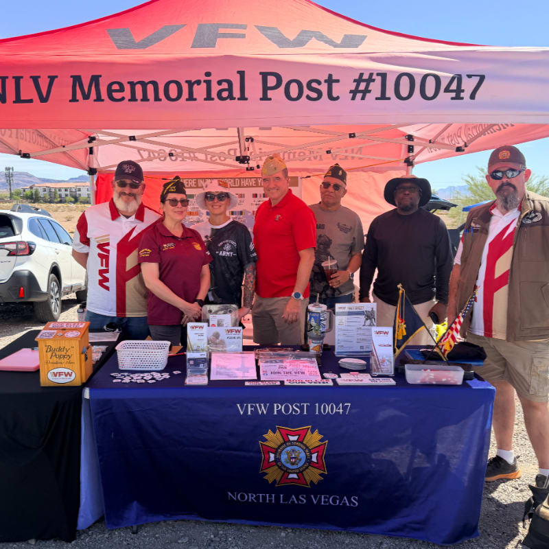 A group of people stand behind a VFW Post #10047 table with promotional materials, under a red tent with the VFW logo, in front of a desert landscape.