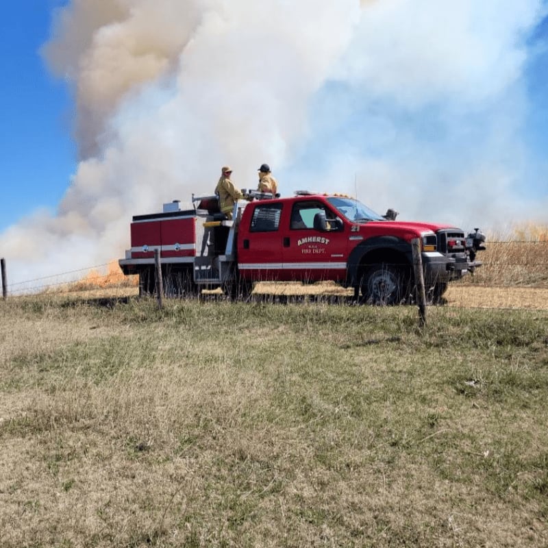 Two firefighters stand on a red fire truck in the foreground, with a large plume of smoke rising from a field in the background under a clear blue sky.