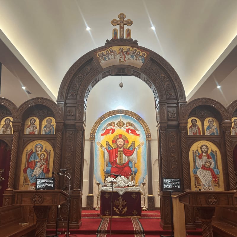 In the foreground, wooden pulpits and a red carpet lead to an altar adorned with religious icons, while the background features a large, ornate icon of Christ framed by arched wooden structures and painted murals.