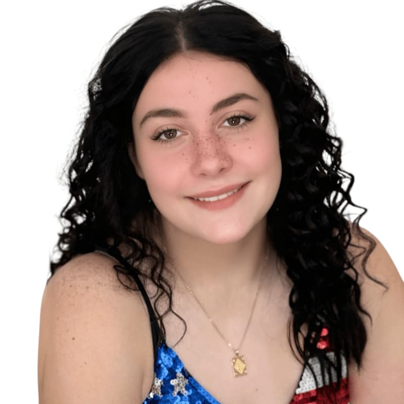 A young woman with dark curly hair and freckles smiles at the camera, wearing a sequined top against a plain white background.