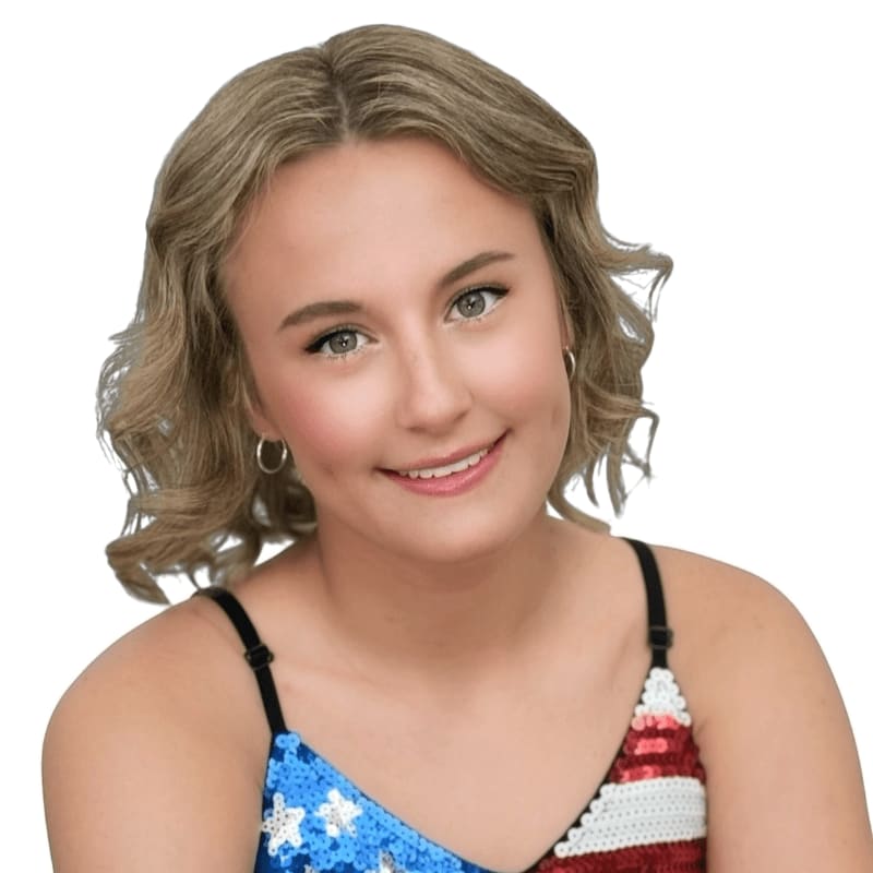 A young woman with blonde, curly hair and a dimpled smile wears a sparkly, American flag-themed top against a plain white background.