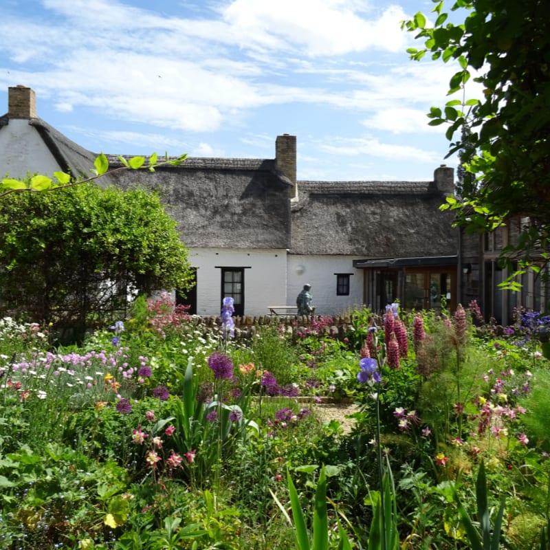 A vibrant, blooming garden fills the foreground, leading to a charming thatched-roof cottage with a person sitting on a bench in the background.