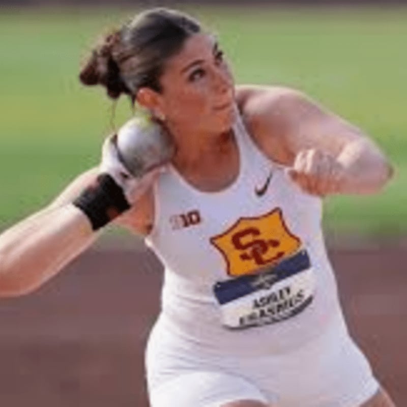 A female athlete in a white uniform prepares to throw a shot put, with a blurred track and field in the background.