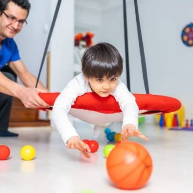 A young child on a red swing reaches for colorful balls on the floor while a man stands behind them in a brightly lit room.