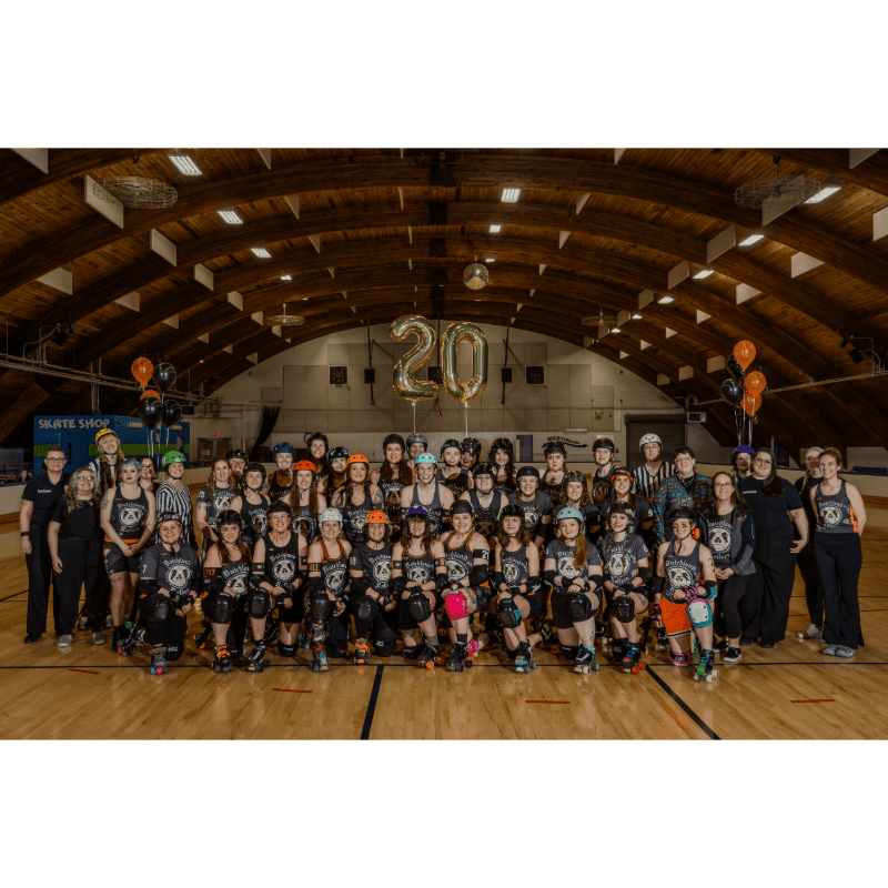 A large group of roller derby skaters in uniform pose for a photo in a gymnasium with large "20" balloons and orange and black balloons in the background.