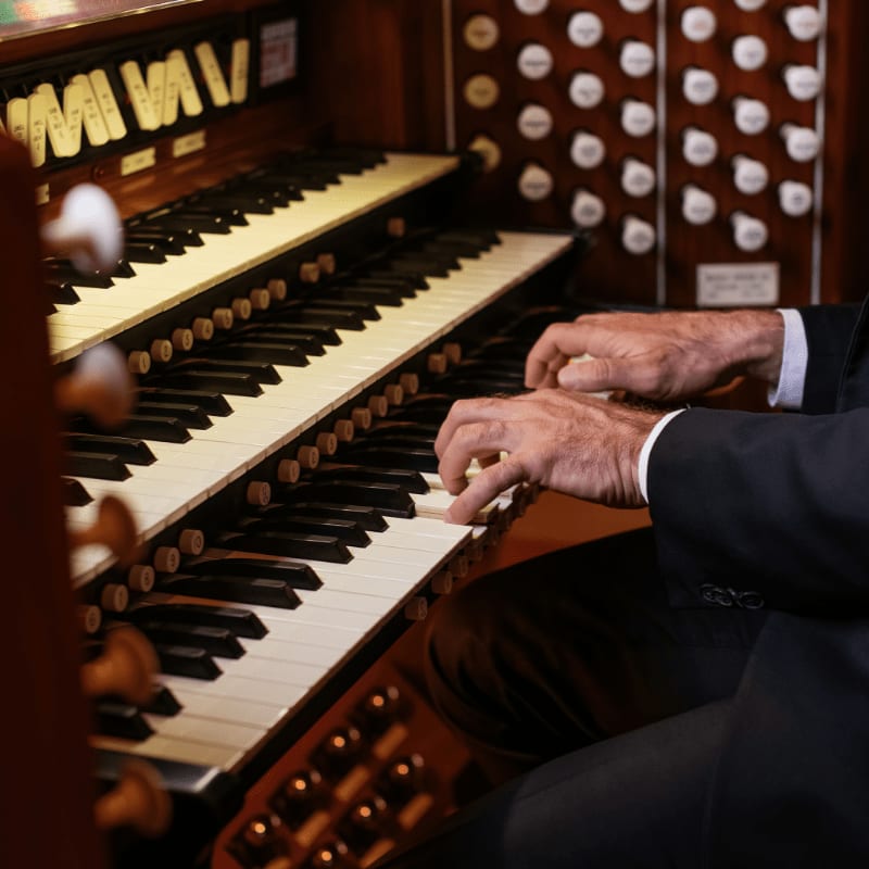 Hands play a pipe organ with its intricate keyboards in the foreground and a wall of pipes in the background.