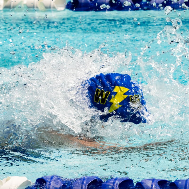 A swimmer in a blue cap with a yellow lightning bolt splashes through the water in a pool with lane markers.