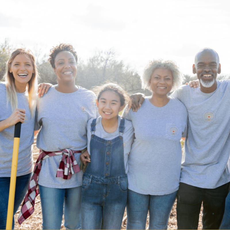 A diverse group of five people, including a young girl in overalls, stand together outdoors with arms around each other, while trees and a bright sky form the background.