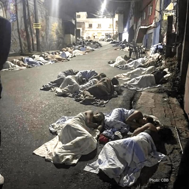 People sleep on the asphalt of a narrow street lined with graffiti-covered walls and buildings in the background.