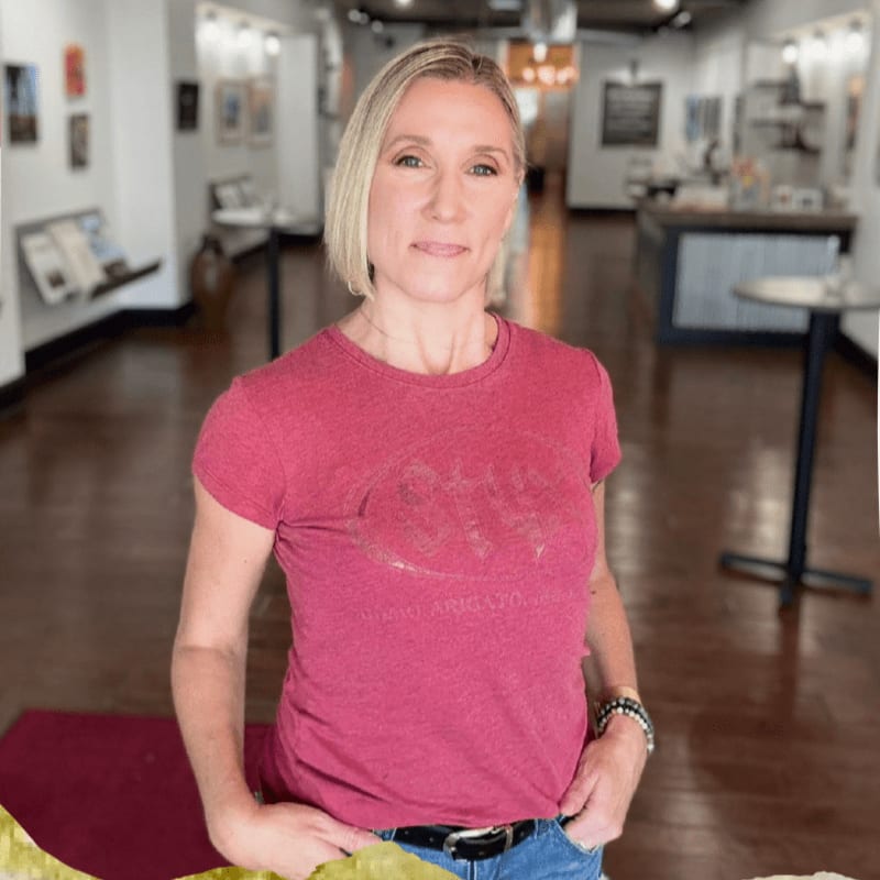 A woman in a maroon t-shirt and jeans stands in the foreground of an art gallery with artwork displayed on the walls in the background.
