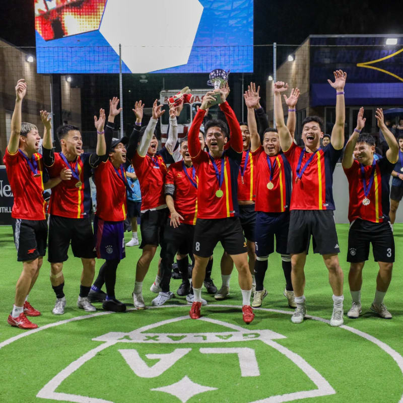 A group of jubilant soccer players in red and yellow jerseys celebrate with a trophy on a turf field with the LA Galaxy logo, while a large screen displays a colorful image in the background.