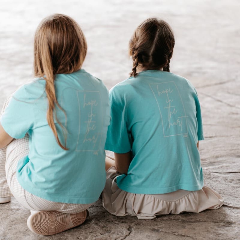 Two girls in matching teal shirts with a white graphic sit on a textured stone floor, with their backs to the camera.