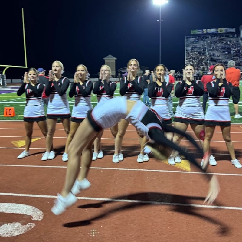 A cheerleader performs a backflip on a football field in front of a line of clapping teammates, with stadium lights and bleachers visible in the background.