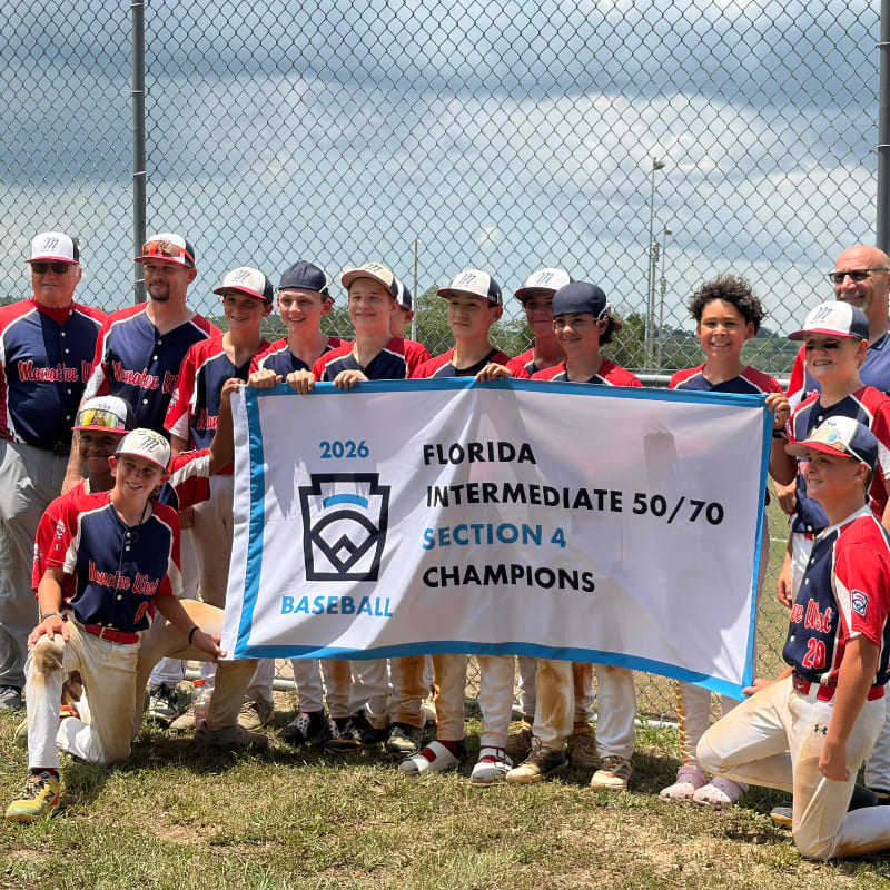 A group of young baseball players in uniform pose with a championship banner in front of a chain-link fence and a cloudy sky.