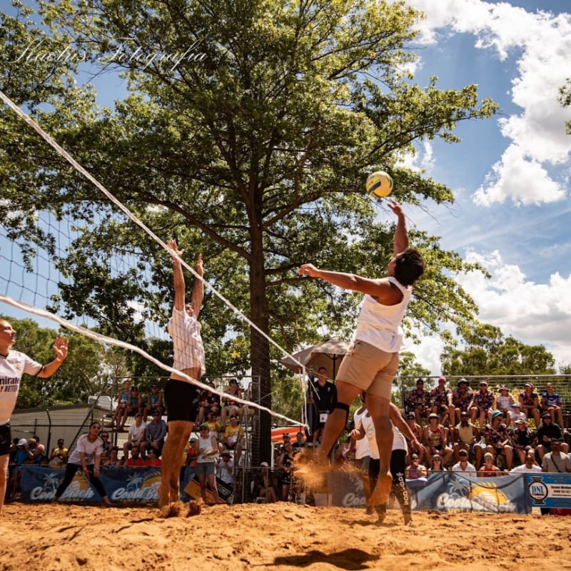 In the foreground, a beach volleyball player jumps to hit the ball over the net, while the background shows a crowd watching the game under a large tree.