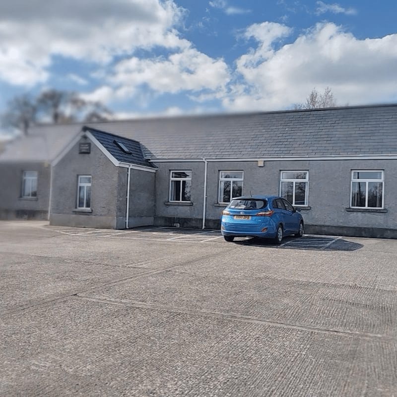 A blue car is parked in a concrete lot in front of a gray building with a slate roof under a cloudy sky.