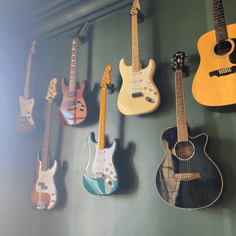 Several electric and acoustic guitars hang on a dark green wall, with a bright light illuminating the left side of the foreground.