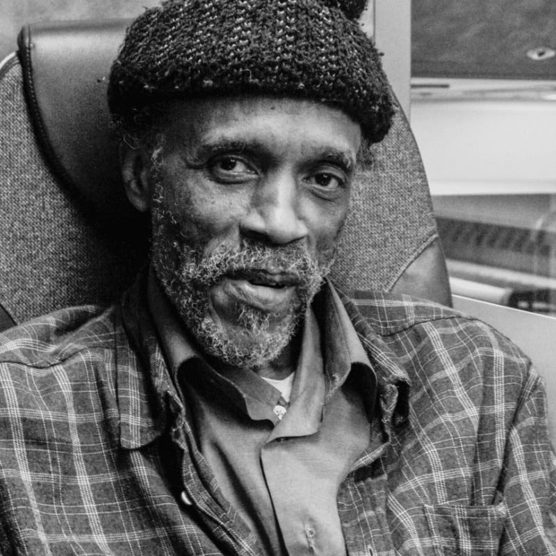 A man in a plaid shirt and cap sits in a chair with a blurred background.