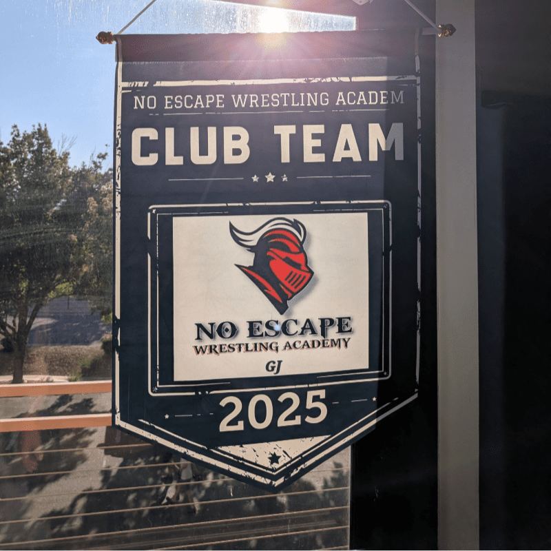 A dark banner for "No Escape Wrestling Academy Club Team 2025" with a knight's helmet logo hangs in front of a sunlit window with trees visible outside.