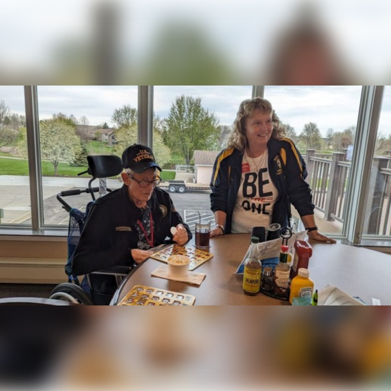 A person in a wheelchair eats at a table with condiments and drinks, while another person stands behind them, with a blurred background of trees and a building.
