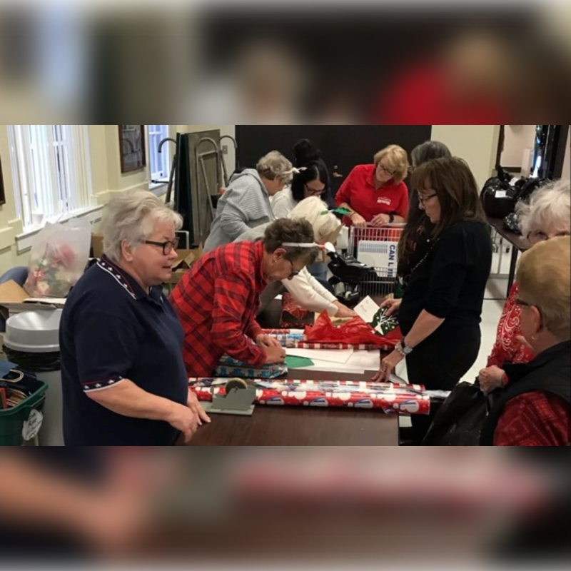In the foreground, a woman in a blue shirt looks to her left, while in the background, several people are gathered around tables, wrapping gifts.
