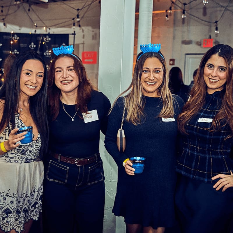 Four smiling women in the foreground wear blue headbands with small bowls, while string lights and a blurred background suggest a festive indoor event.