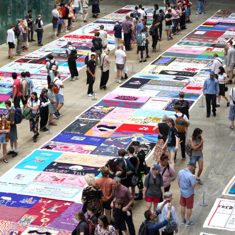 People stand around a long, colorful quilt laid out on the floor in a large, open hall.