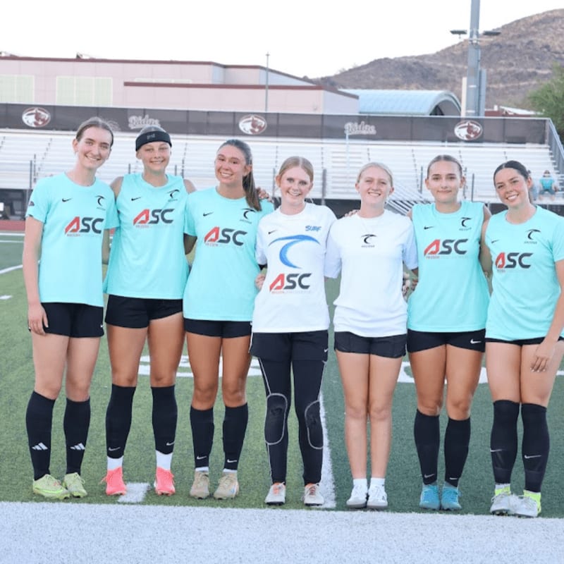 Six young women in athletic wear stand on a green field in front of bleachers and a desert landscape.