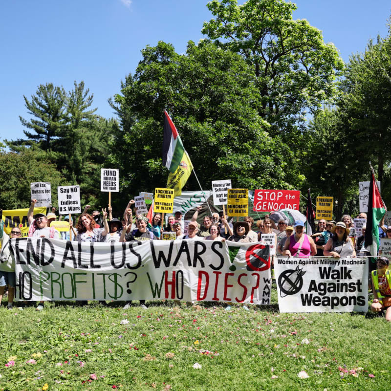 A crowd of protestors holding signs and banners advocating for peace stands in a grassy park with trees in the background.