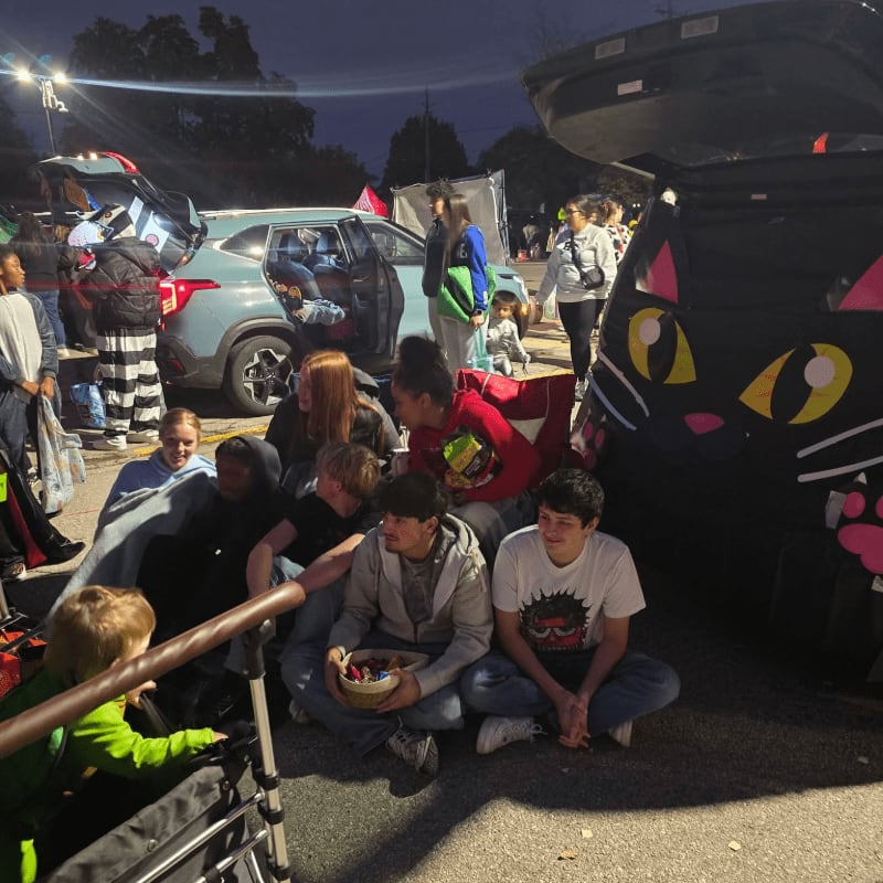 Children and adults gather around decorated car trunks at night, with a large black cat decoration in the foreground and cars and lights in the background.