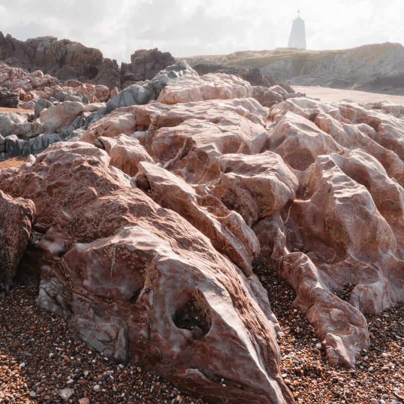 Rough, reddish-brown rocks fill the foreground of a rocky beach, with a white lighthouse visible on a distant grassy hill under a cloudy sky.