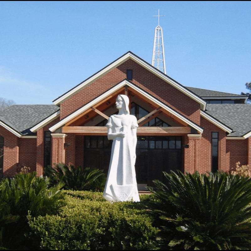 A white statue stands in front of lush green bushes, with a brick church featuring a prominent steeple in the background.
