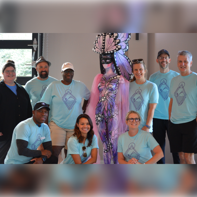 A group of people in light blue t-shirts pose in front of a colorful mannequin with butterfly wings, with a blurred background of a room.
