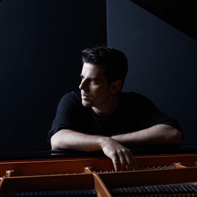 A man with his arms resting on a piano looks off to the side against a dark background.