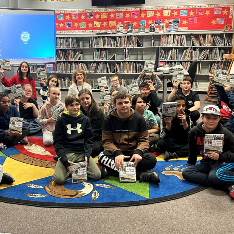 Children and adults sit on a colorful rug in front of a bookshelf filled with books, holding up copies of a book.