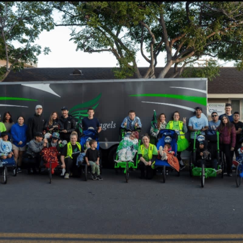 A group of people, some in wheelchairs, pose in front of a large trailer with green accents, with houses and trees in the background.
