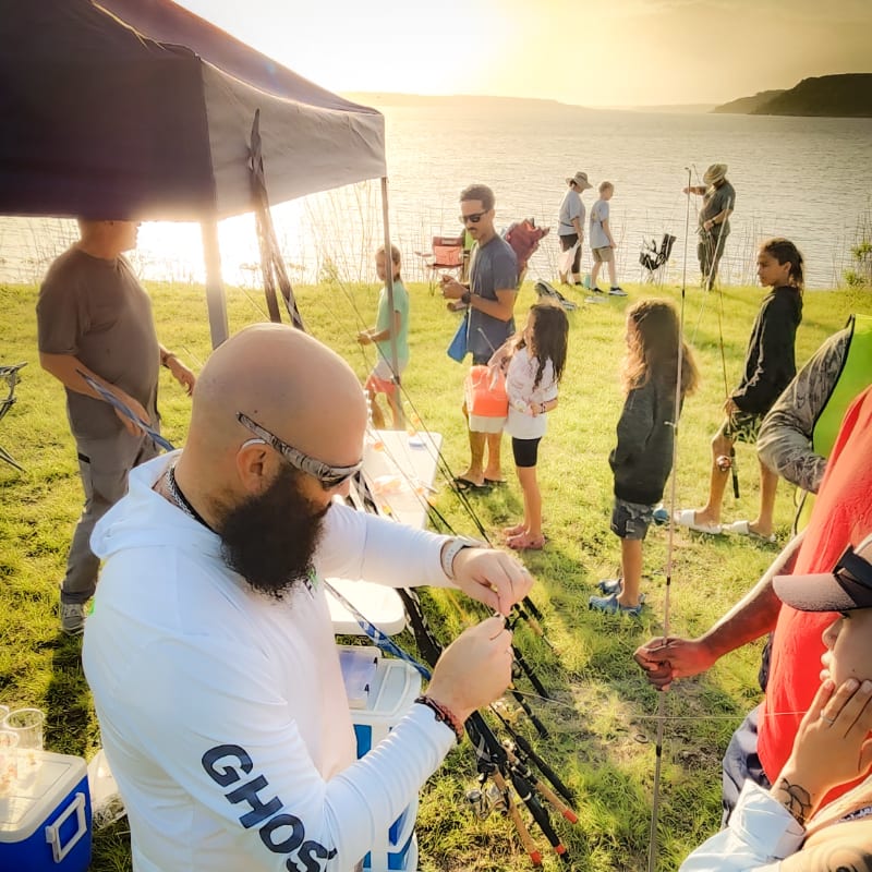 A group of people, including children, are gathered for fishing under a tent in the foreground, with a calm lake and sunset in the background.