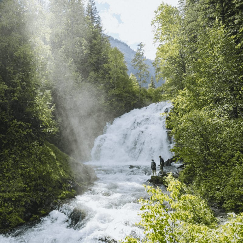 Two people stand on rocks in the foreground of a powerful waterfall cascading down a lush, green mountainside.