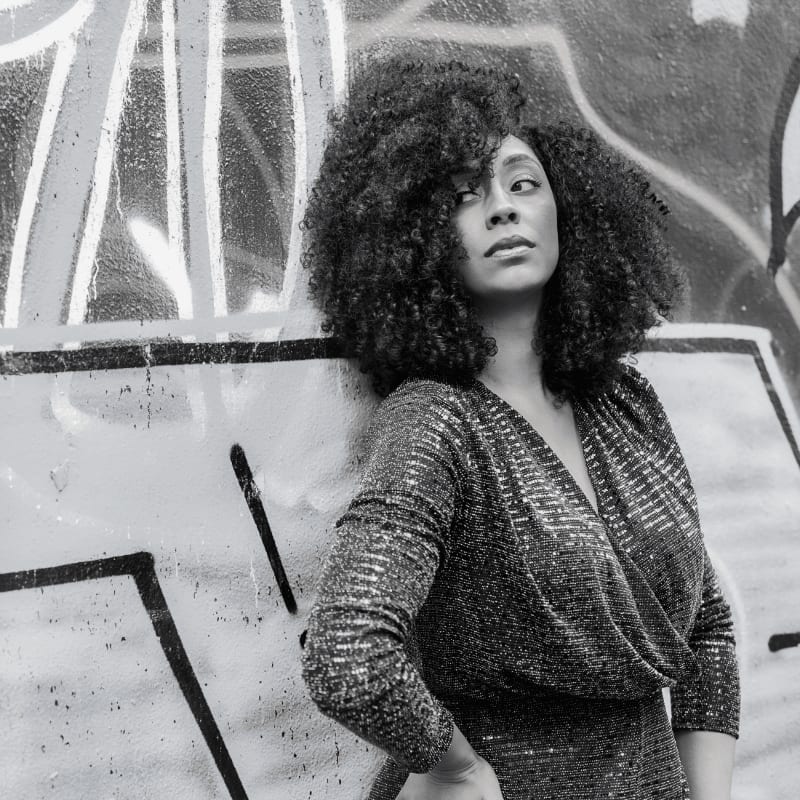 A woman with voluminous curly hair leans against a graffiti-covered wall.