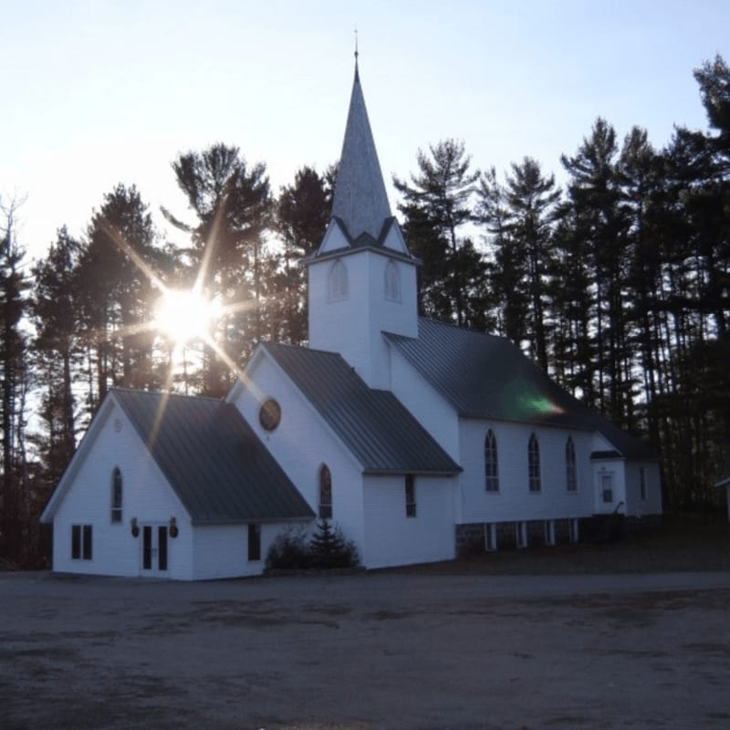 A white church with a tall steeple stands in the foreground, silhouetted against a backdrop of dark pine trees and a bright sunburst.