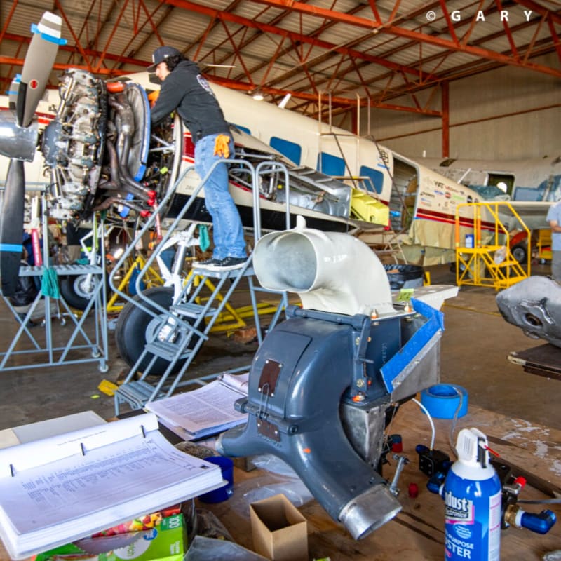 A mechanic works on an airplane engine in a hangar, with various tools and parts scattered on the floor in the foreground.