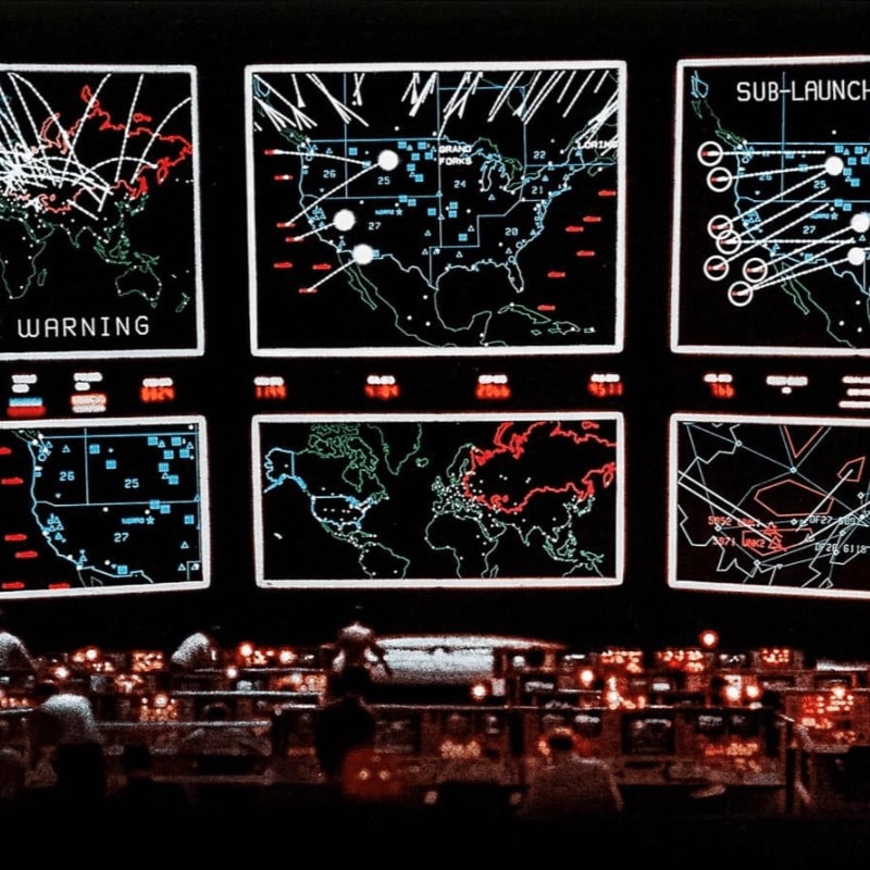 In the foreground, silhouetted figures are seated at consoles, while the background displays a grid of six screens showing maps with various data overlays, including missile trajectories and warning indicators.