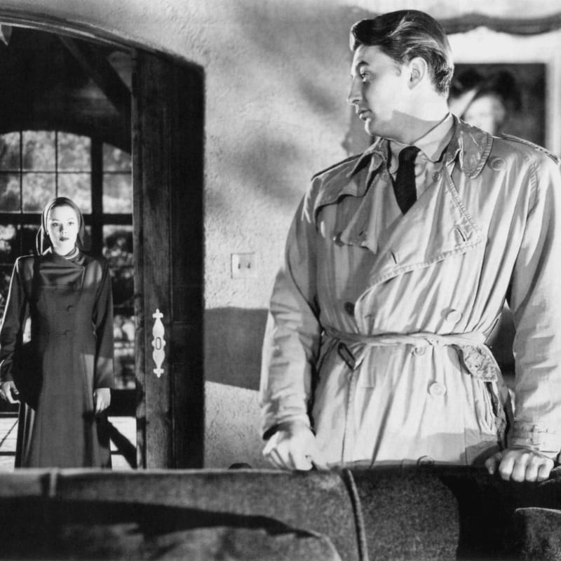 A man in a trench coat stands in the foreground, looking towards a woman in a dark dress standing in a doorway in the background.