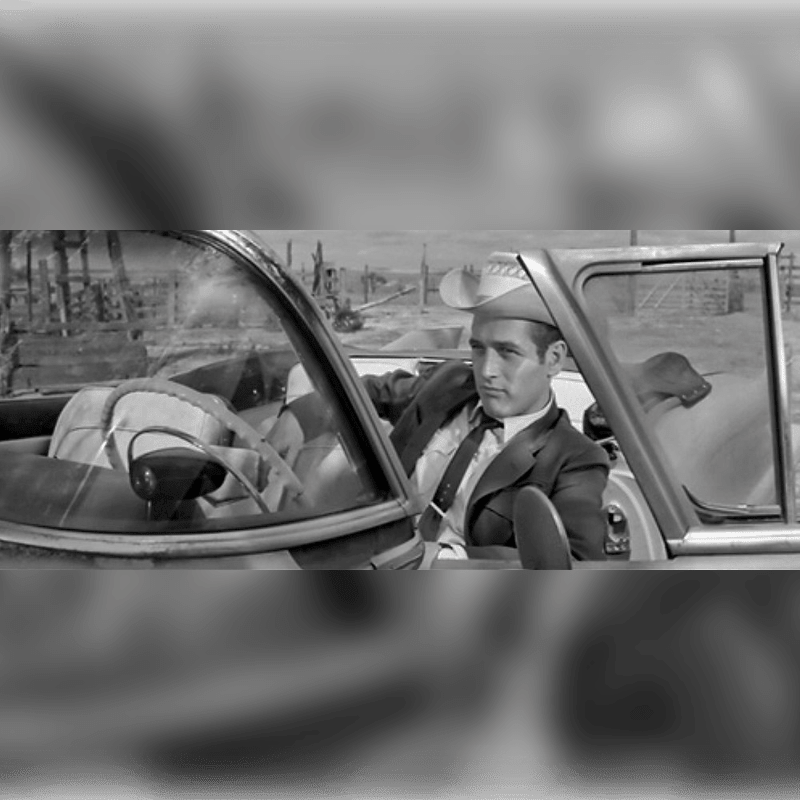 A man in a cowboy hat and suit sits in a convertible with luggage in the passenger seat, while a desolate, arid landscape stretches out behind him.