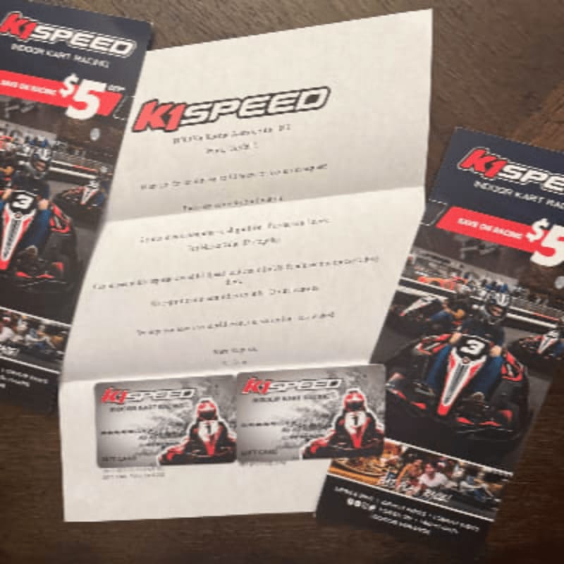 Two gift cards and a folded letter from K1 Speed are laid out on a wooden surface, with flyers showing go-karts in the background.
