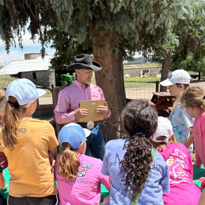 A man in a cowboy hat and pink shirt stands in front of a group of children gathered under a tree, with farm buildings and a fence in the background.