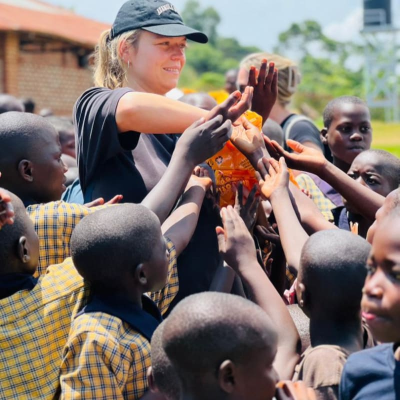 A young woman in a black cap and shirt distributes items to a crowd of eager children in yellow and brown plaid uniforms, with a blurred background of buildings and greenery.