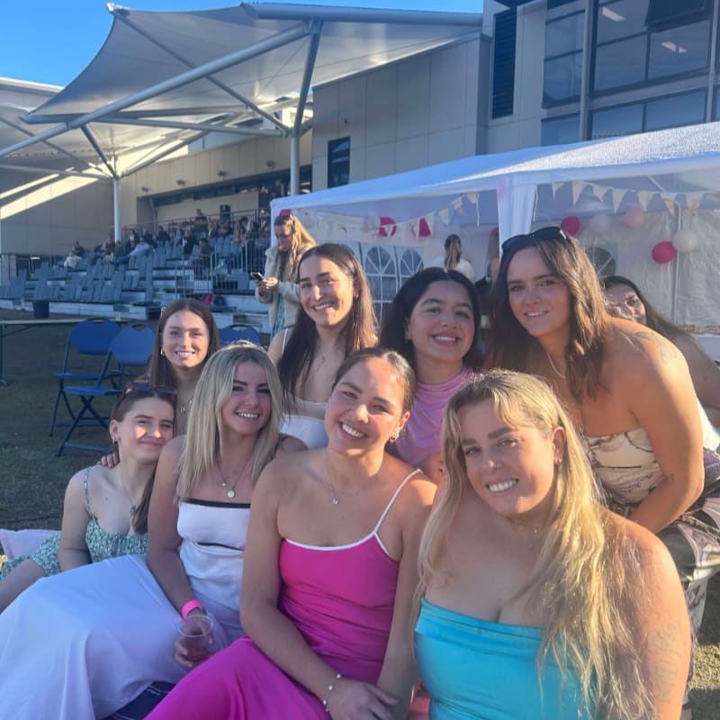 A group of smiling young women pose for a photo in the foreground, with a seated audience and a white tent in the background.