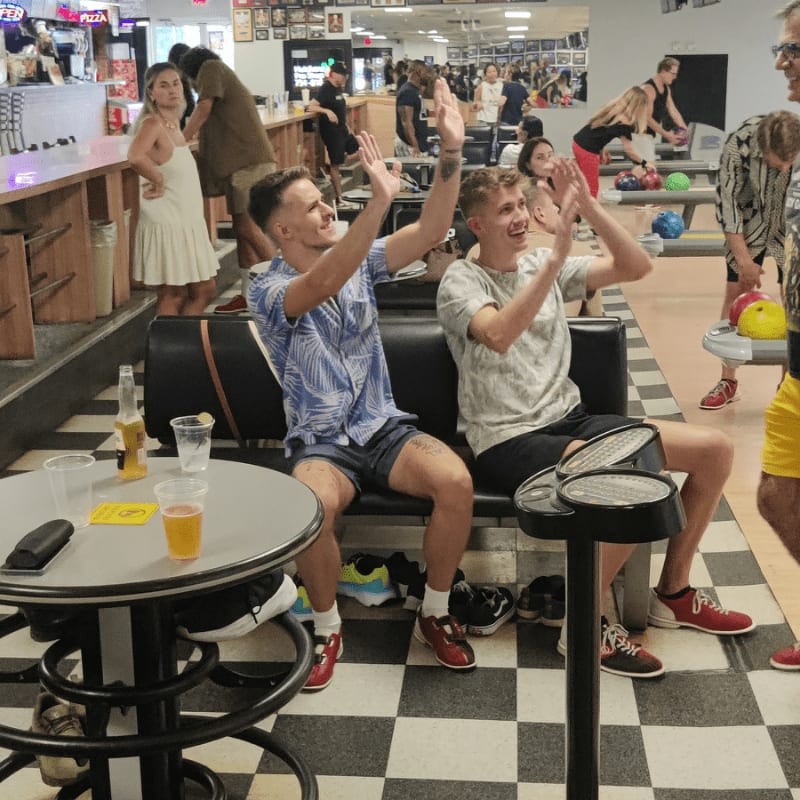 Two young men in the foreground high-five while sitting on a bench in a bowling alley, with other patrons and bowling lanes visible in the background.